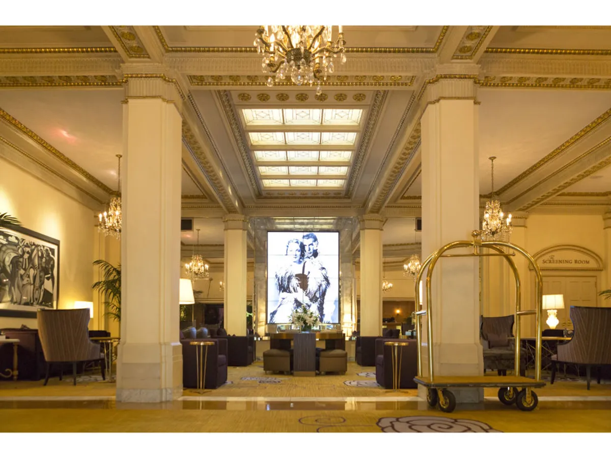 A luxurious hotel lobby interior with grand chandeliers, large artwork, multiple seating areas, and a welcoming lobby cart. The scene is bustling with social activity, reflecting a comfortable yet sophisticated gathering space.