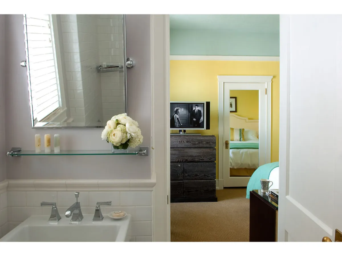 A serene bathroom scene featuring a well-lit sink area, a stylish mirror, a vase of fresh flowers, and hints of a yellow accent wall. The presence of a television visible from the bathroom area hints at a space designed for relaxation and a luxurious, self-care experience.