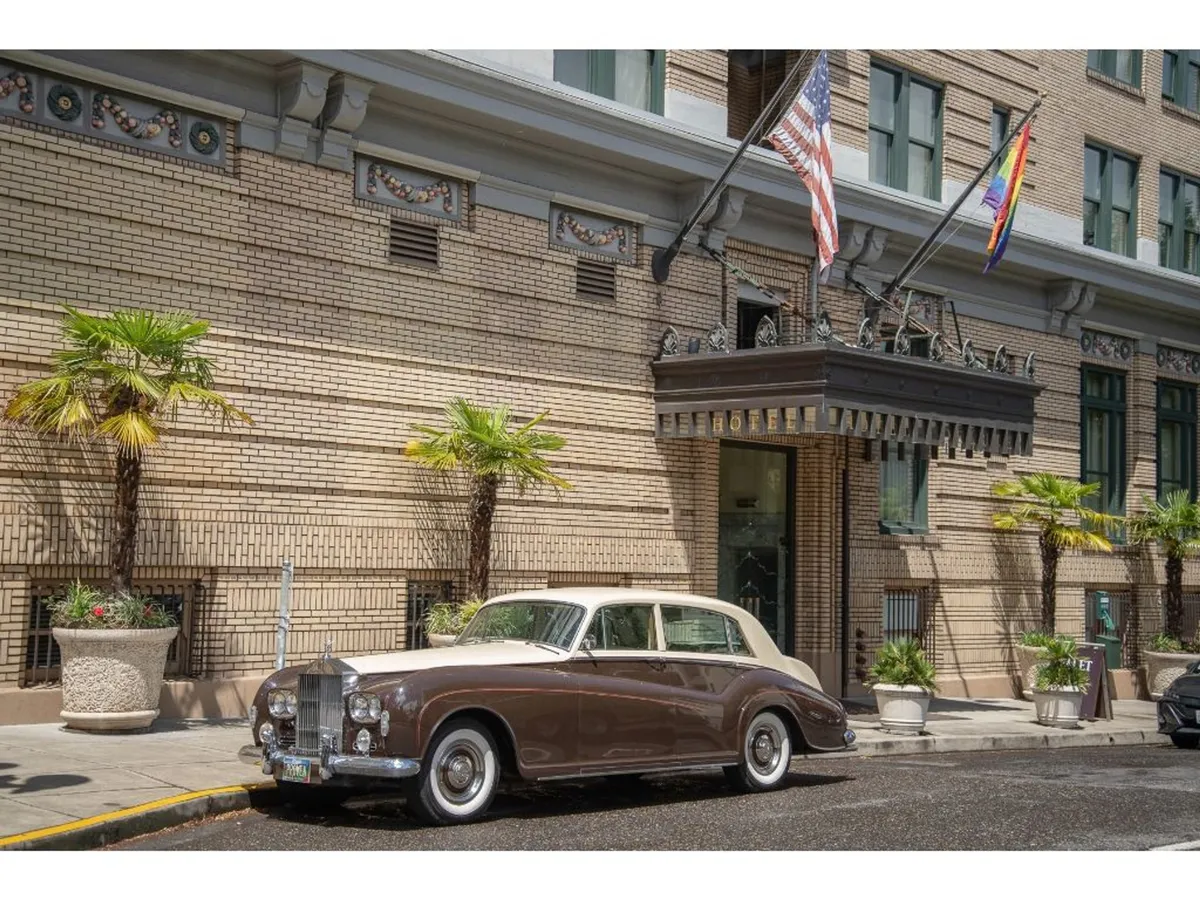 The image showcases the grand exterior of Hotel Deluxe, featuring a classic car parked in front, palm trees, fluttering flags, and the elegant, inviting facade. The scene is lively, with guests arriving and a sense of anticipation for upcoming events.