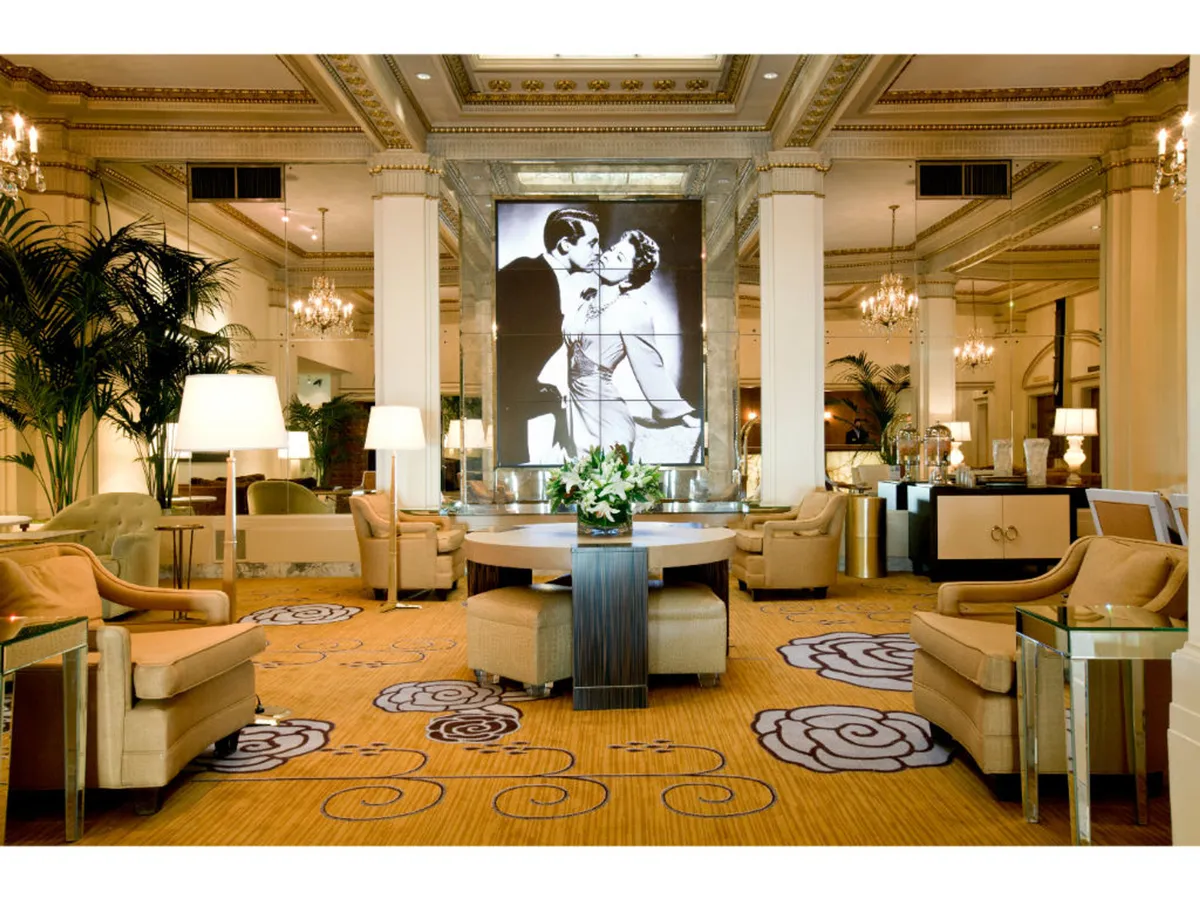 A luxurious interior scene featuring a large, romantic artwork of a couple kissing, a grand table with a dramatic floral arrangement, and plush seating areas arranged for social gatherings or receptions. The atmosphere is both sophisticated and intimate, perfect for special celebrations or casual meetups.