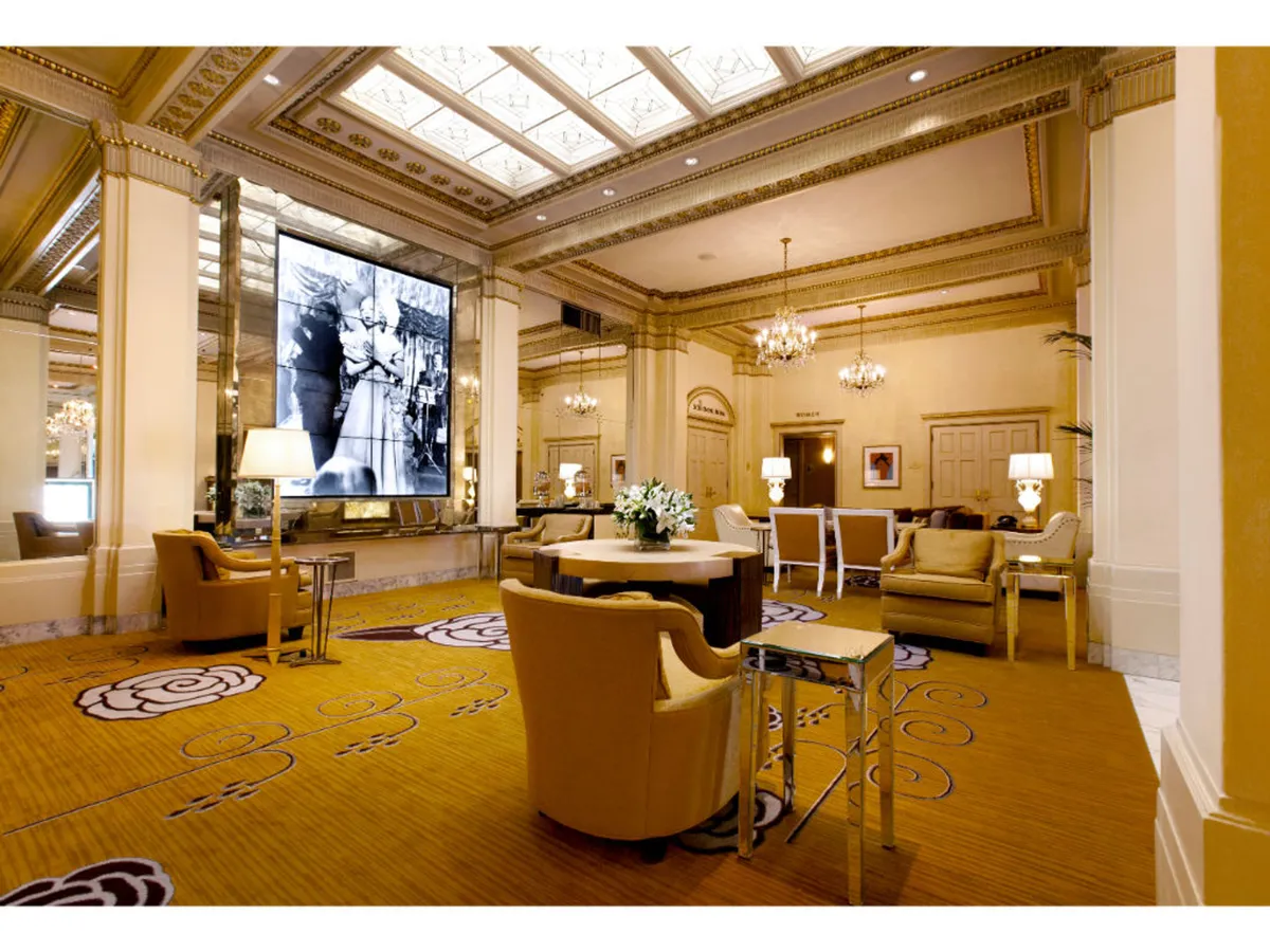 A sophisticated hotel lounge with a prominent large digital screen displaying a historic black and white photograph, a central round table with a floral arrangement, elegant chandeliers, and plush seating clusters. The space exudes cinematic charm and is set up for social gatherings, business meetings, or private events.
