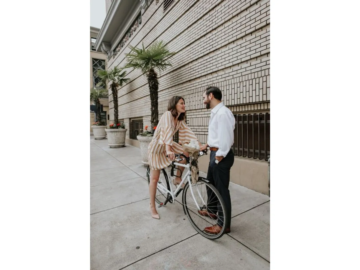 An inviting exterior scene featuring a couple enjoying a casual conversation beside a bicycle, with potted plants and the hotel's building facade in the background. The setting radiates a relaxed urban vibe, with the bike subtly referencing the hotel's complimentary bicycles and the active, walkable downtown location.