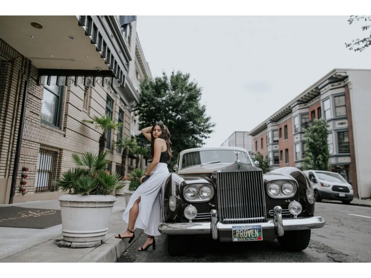 An elegant exterior shot featuring a vintage car parked in front of the building's facade, with a model in a white dress and lush potted plants framing the scene. The image captures a sense of urban chic and romantic exploration, suggesting the excitement of stepping out for stylish adventures downtown.