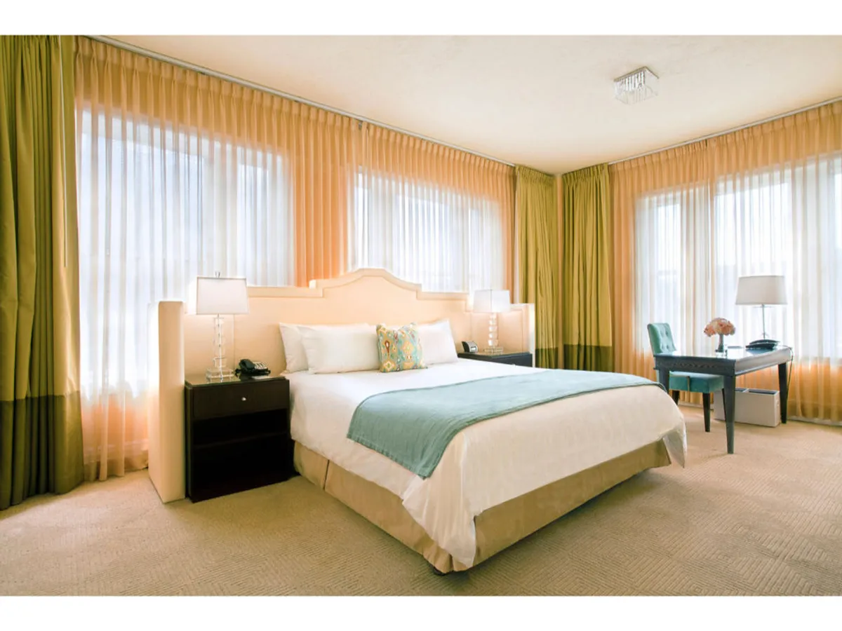 The image showcases a sunlit studio suite with a plush king bed adorned with decorative pillows, oversized windows draped in sheer curtains, and an elegant desk topped with a lamp and fresh flowers. The tranquil setting is enhanced by classic crown moldings and soft, natural light flooding the space, creating an inviting corner for relaxation or productivity.
